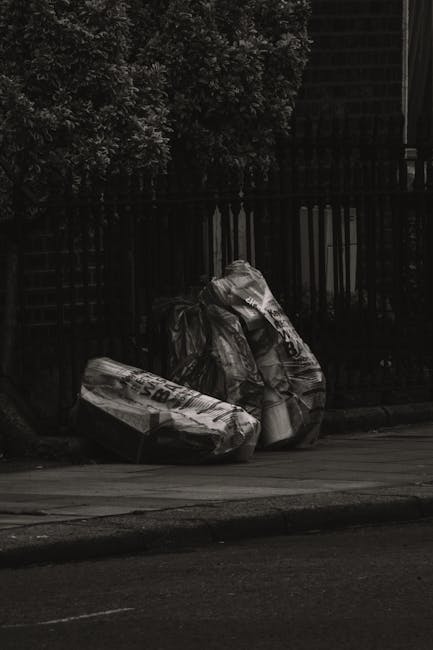 A collection of large, crumpled black plastic rubbish bags resting on a paved sidewalk next to a curb. The bags appear to contain general waste and are secured with visible knots, with some displaying partially torn or creased surfaces. They are positioned close to a black metal fence with vertical bars, which runs parallel to the street and separates the sidewalk from a background area that includes tall, leafy trees providing partial shade. The environment suggests an urban setting, possibly a designated collection point for waste disposal. The scene is lit with natural daylight, casting soft shadows, and the overall atmosphere is calm, with no visible activity or people in the immediate vicinity. This image illustrates a typical scenario where private waste handling services might be employed to manage rubbish removal, emphasizing the importance of proper waste collection for maintaining cleanliness in residential or public spaces.