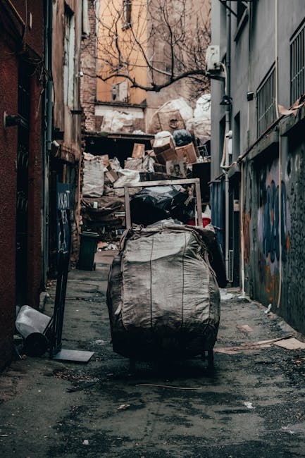 A narrow urban alleyway filled with overflowing rubbish and debris. In the foreground, a large, grey, weathered bag made of heavy-duty material is placed on the uneven, dark asphalt ground, which shows signs of staining and dirt. Behind the bag, there are stacked cardboard boxes, plastic bags, and other waste materials piled against a backdrop of old brick buildings and a metal fence. The alley is flanked by walls covered in graffiti and peeling paint, with some bare brick visible near the top. Bare tree branches extend into the scene from the upper left, indicating an outdoor, city-centre environment. The lighting is natural, likely overcast, casting a muted tone over the scene, emphasizing the cluttered and neglected appearance typical of private waste storage in urban alleys. This setting illustrates a situation where independent rubbish collection or disposal services, such as those offered by Rubbish Removal Streatham, might be needed to clear accumulated waste efficiently and avoid issues associated with unmanaged rubbish accumulation in urban areas.