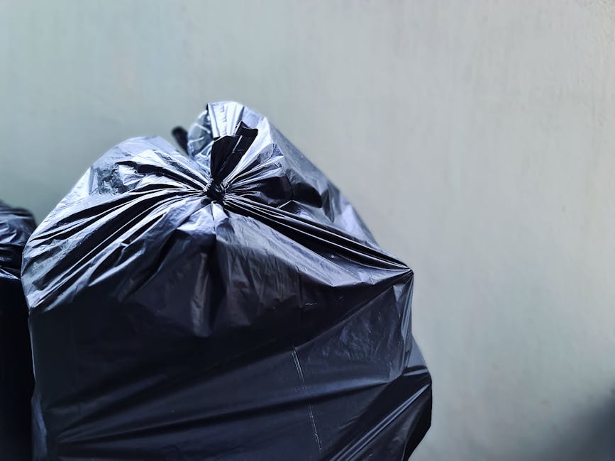 A collection of large, crumpled black plastic rubbish bags resting on a paved sidewalk next to a curb. The bags appear to contain general waste and are secured with visible knots, with some displaying partially torn or creased surfaces. They are positioned close to a black metal fence with vertical bars, which runs parallel to the street and separates the sidewalk from a background area that includes tall, leafy trees providing partial shade. The environment suggests an urban setting, possibly a designated collection point for waste disposal. The scene is lit with natural daylight, casting soft shadows, and the overall atmosphere is calm, with no visible activity or people in the immediate vicinity. This image illustrates a typical scenario where private waste handling services might be employed to manage rubbish removal, emphasizing the importance of proper waste collection for maintaining cleanliness in residential or public spaces.