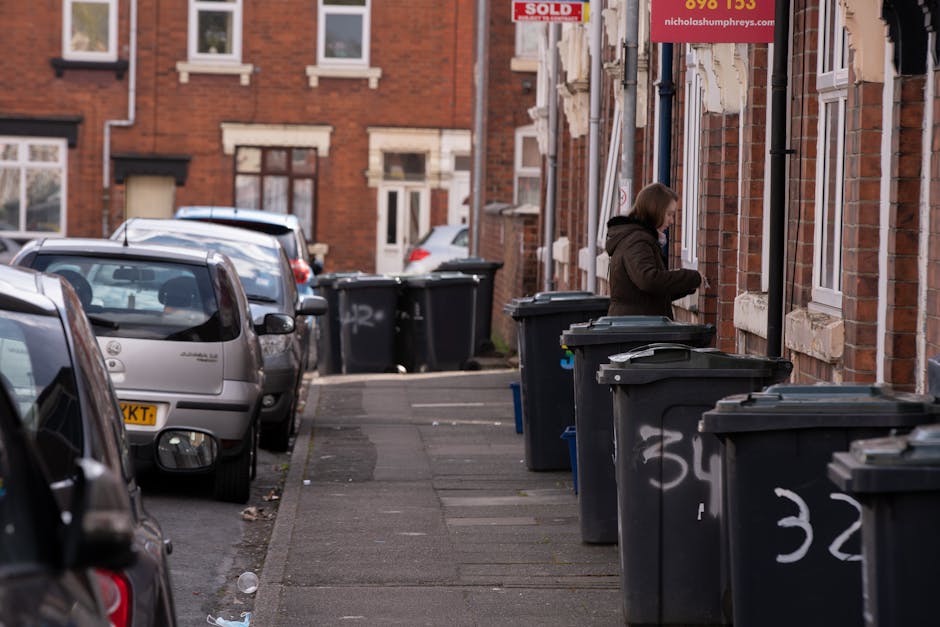 A residential street scene features a row of dark grey wheelie bins lined up along the curbside, each with closed lids and some marked with white spray-painted numbers. A woman with shoulder-length hair, dressed in a brown coat, is standing next to one of the bins, seemingly preparing to open it or discard waste. The background displays red brick terraced houses with white-framed windows and a red 'SOLD' sign partially visible on a building facade, indicating these are private properties. Parked cars of various models and colors, including silver, grey, and dark tones, are lined along the street, occupying designated parking spaces adjacent to the sidewalk. The sidewalk itself is paved with concrete slabs, with some litter visible near the curb. The lighting appears to be natural daylight, providing a neutral and clear view of the scene's details. This image highlights typical urban waste management practices, with private collection bins positioned outside homes, aligning with alternative rubbish removal services such as same-day or on-site clearance managed by companies like Rubbish Removal Streatham.