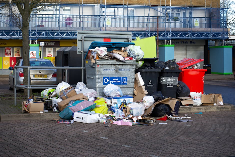 A collection of overflowing rubbish bins and scattered waste on a paved sidewalk in front of a commercial building. The waste includes black and red garbage bags, flattened cardboard boxes, paper and plastic packaging, and various discarded items such as cardboard containers and plastic wrappers. The bins are made of metal and plastic, with the central one labeled for mixed paper and cardboard, showing signs of being overstuffed with items spilling out onto the ground. Surrounding the waste are parked cars, including a grey vehicle on the left, and a metal railing separating the rubbish area from the parking spaces. In the background, there is a building under renovation or construction, covered with blue scaffolding and protective netting, with some signage visible. The scene appears to depict a typical instance of unmanaged waste accumulation, which could be managed more effectively through services such as those provided by Rubbish Removal Streatham, focusing on private or alternative rubbish collection methods.