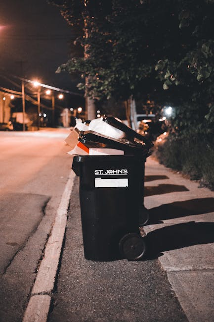 A residential street scene features a row of dark grey wheelie bins lined up along the curbside, each with closed lids and some marked with white spray-painted numbers. A woman with shoulder-length hair, dressed in a brown coat, is standing next to one of the bins, seemingly preparing to open it or discard waste. The background displays red brick terraced houses with white-framed windows and a red 'SOLD' sign partially visible on a building facade, indicating these are private properties. Parked cars of various models and colors, including silver, grey, and dark tones, are lined along the street, occupying designated parking spaces adjacent to the sidewalk. The sidewalk itself is paved with concrete slabs, with some litter visible near the curb. The lighting appears to be natural daylight, providing a neutral and clear view of the scene's details. This image highlights typical urban waste management practices, with private collection bins positioned outside homes, aligning with alternative rubbish removal services such as same-day or on-site clearance managed by companies like Rubbish Removal Streatham.