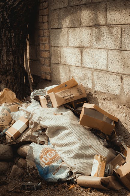 A collection of discarded cardboard boxes, some flattened and others partially intact, are piled against a rough, textured brick wall outdoors. The boxes vary in color from brown to lighter shades, with some printed labels visible on their surfaces. Intermixed with the boxes are crumpled brown paper bags and a large, woven fabric sack that appears to contain additional waste. The pile is situated on uneven ground with small stones and dirt visible beneath it. To the left, part of a tree trunk with dark, rough bark is seen, indicating the waste is in an outdoor area, likely a back alley or side boundary. The lighting is natural, suggesting daylight that illuminates the scene clearly. The scene reflects typical informal rubbish disposal, possibly related to private waste handling services such as Rubbish Removal Streatham, offering an alternative to council collection for the disposal of household or commercial waste.