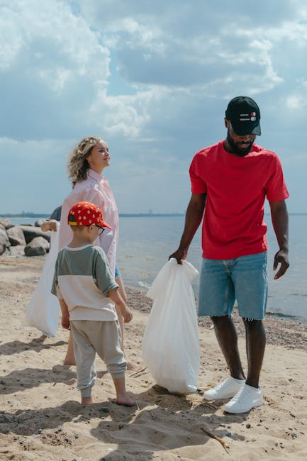 A group of three people, including a man and two children, are seen participating in a beach clean-up activity by the water's edge. The man, dressed in a red T-shirt, light blue denim shorts, and a black cap, is holding a large white rubbish bag while standing on sandy ground. One child, wearing a grey T-shirt, beige pants, and a red cap with white polka dots, is facing toward the water and is holding a piece of trash in their hand. The other child, a girl with curly blonde hair, dressed in a light pink hoodie and blue jeans, stands nearby, appearing to observe the activity. In the background, there are large rocks lining the shoreline, silent waters extending toward the horizon, and a partly cloudy sky overhead. The scene reflects an outdoor effort to clear litter, aligning with rubbish removal services focused on environmental cleanup, particularly in settings such as beaches or natural outdoor areas. Natural lighting highlights the textures of the sand, the fabric of clothing, and the plastic rubbish bags, emphasizing the act of collecting refuse during the day near a body of water.