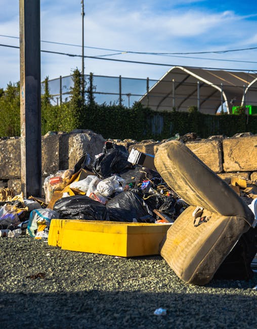 A collection of discarded cardboard boxes, some flattened and others partially intact, are piled against a rough, textured brick wall outdoors. The boxes vary in color from brown to lighter shades, with some printed labels visible on their surfaces. Intermixed with the boxes are crumpled brown paper bags and a large, woven fabric sack that appears to contain additional waste. The pile is situated on uneven ground with small stones and dirt visible beneath it. To the left, part of a tree trunk with dark, rough bark is seen, indicating the waste is in an outdoor area, likely a back alley or side boundary. The lighting is natural, suggesting daylight that illuminates the scene clearly. The scene reflects typical informal rubbish disposal, possibly related to private waste handling services such as Rubbish Removal Streatham, offering an alternative to council collection for the disposal of household or commercial waste.